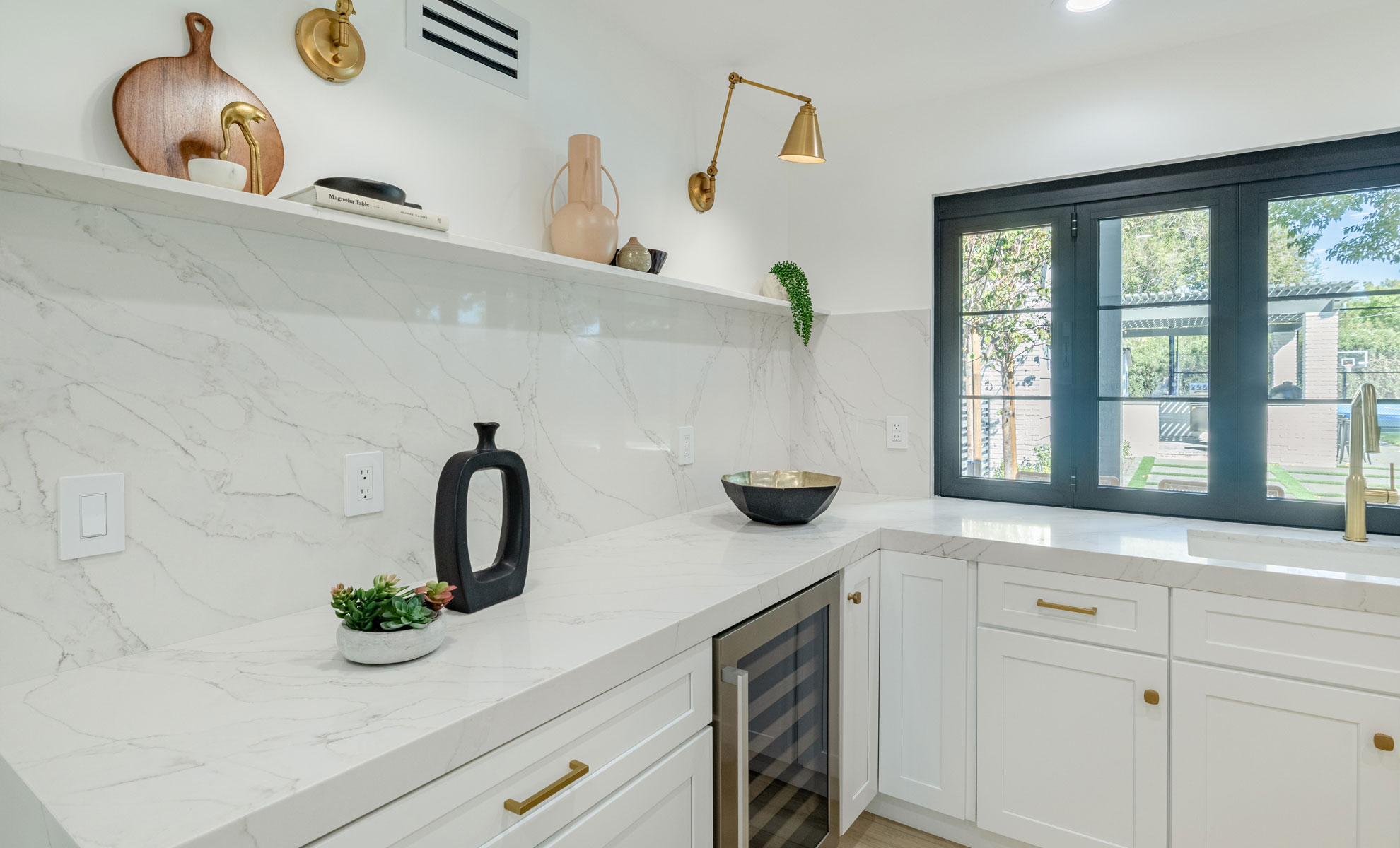 This kitchen features a white quartz countertop and backsplash with veins. A plant, a bowl and a vase are resting on the countertop. A cutting board, a book, a plant and a vase are set on a shelf above the countertop.