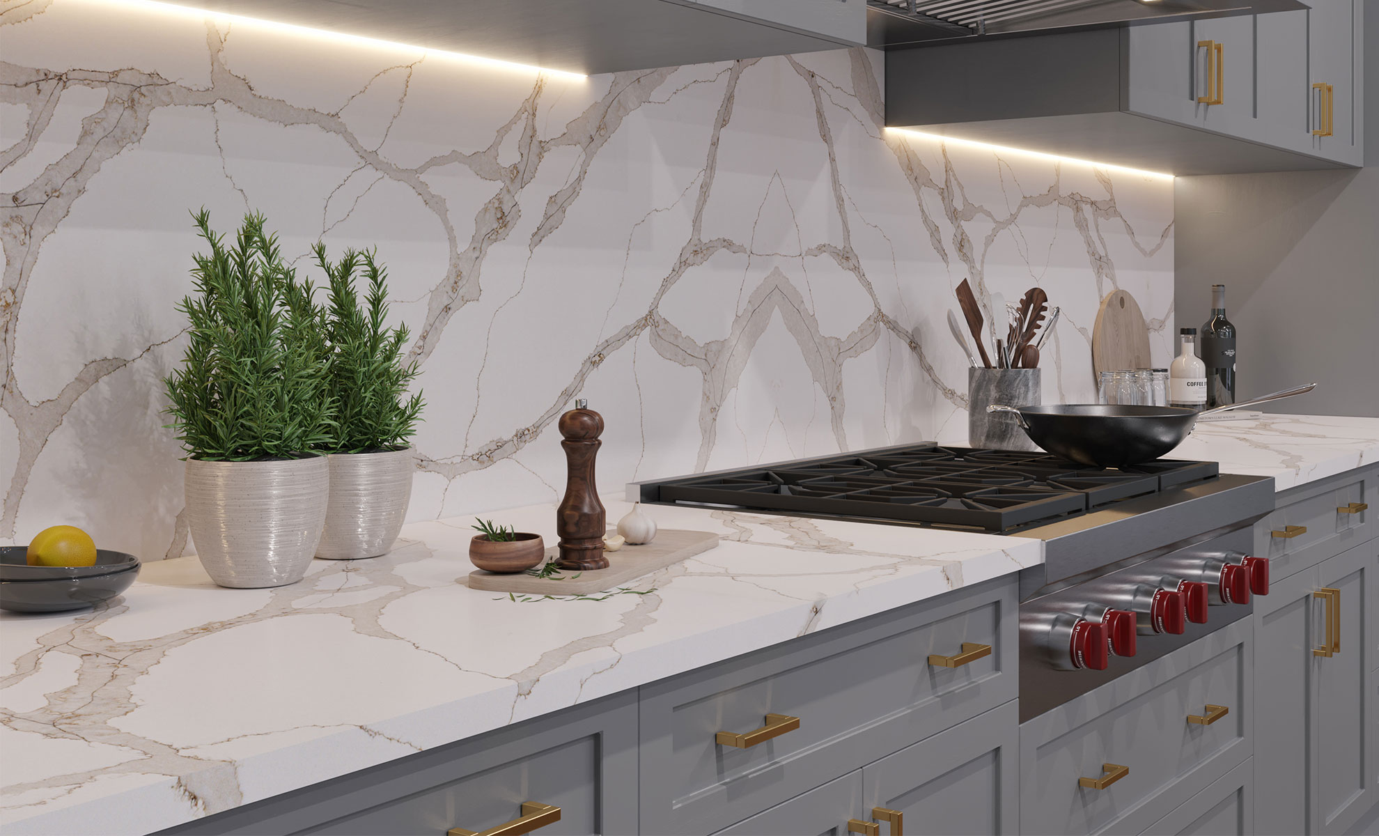 This kitchen features a gorgeous countertop and backsplash. There are grey cabinets with gold hardware incorporated into the design. Various kitchen items, including a pan, a utensil holder, a cutting board, two herb plants and a pepper shaker, are set on the counter.