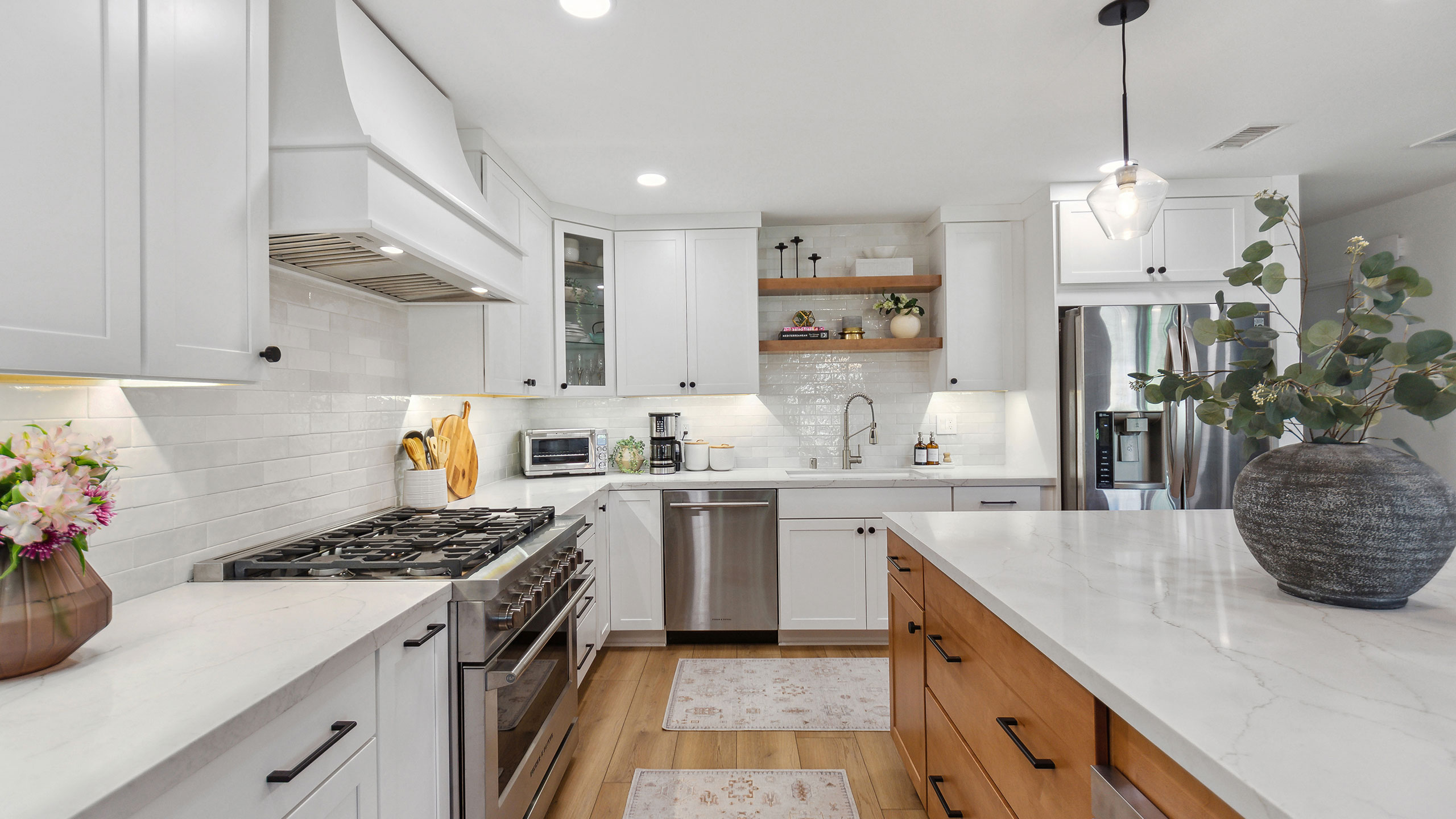 This kitchen features white cabinets and Vadara Ostara Dawn white quartz countertops. The kitchen island has brown wood cabinets and a potted plant above on top of it.