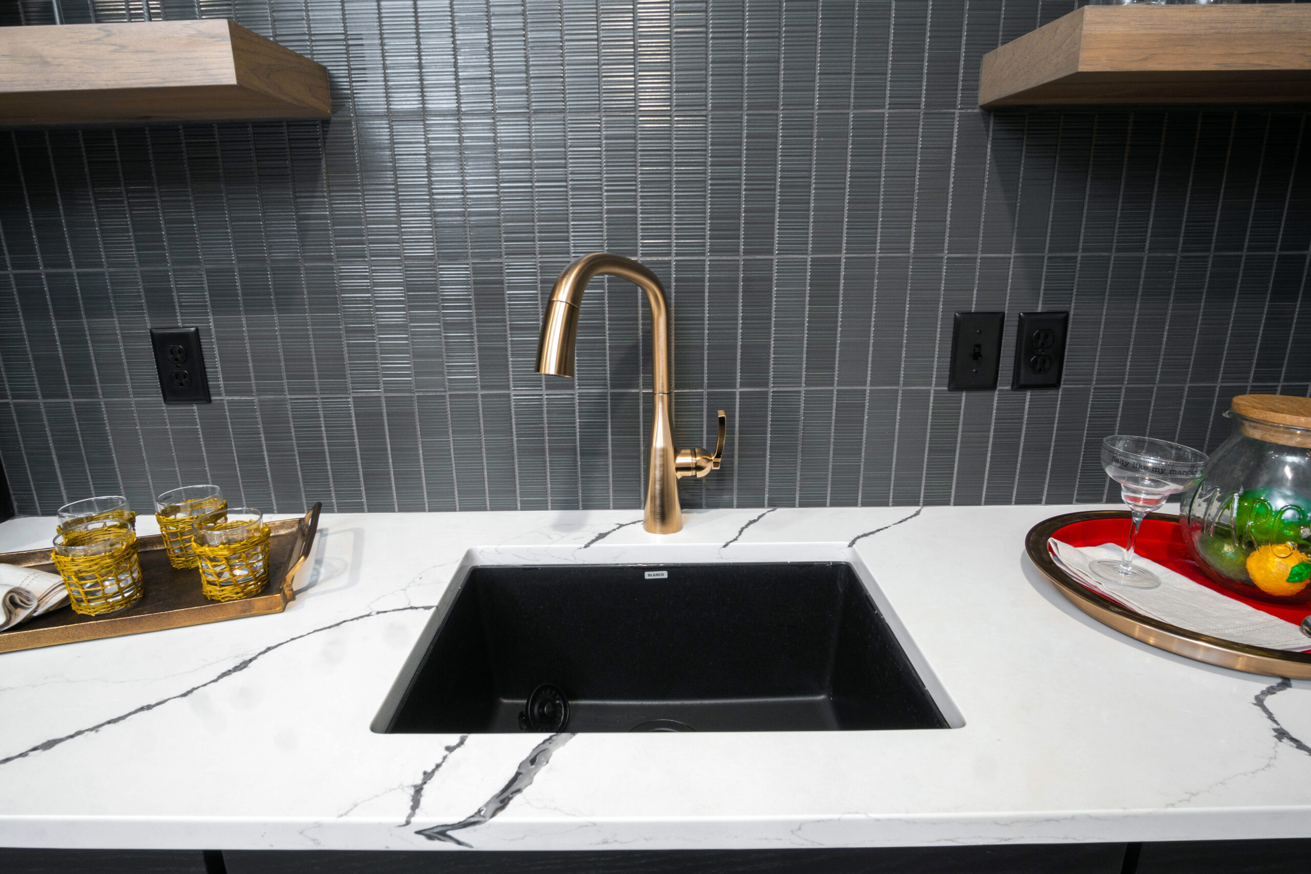This kitchen features a black sink with a gold faucet surrounded by white countertop with black and gray veins. The backsplash features gray tiles.