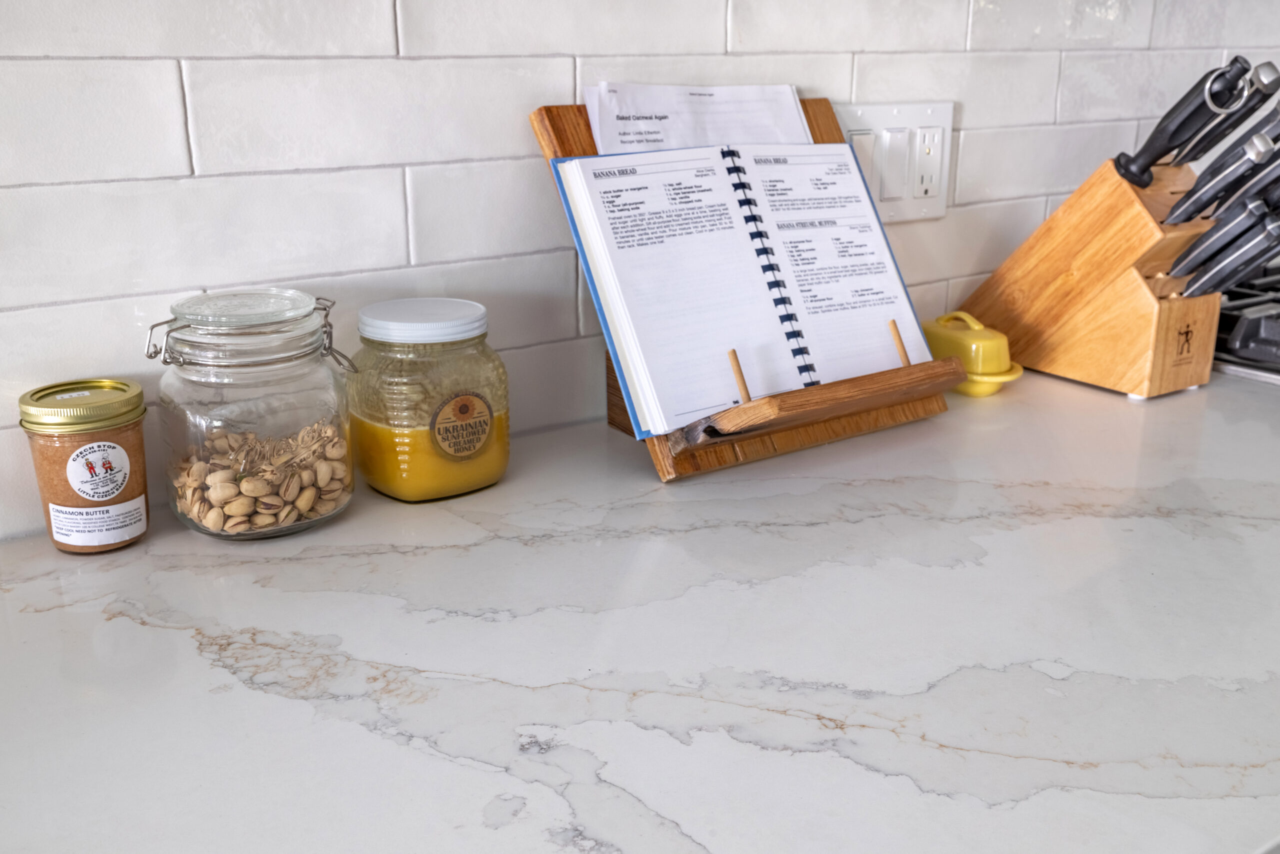 This kitchen features a white countertop with grey and gold veins. There is an open book, jars, a butter holder and a knife set resting on the countertop against the white-tiled backsplash.